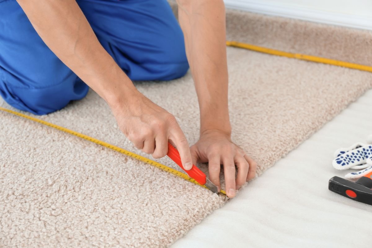 A person using a utility knife to cut carpet while measuring with a yellow tape measure on a soft, textured carpet surface.