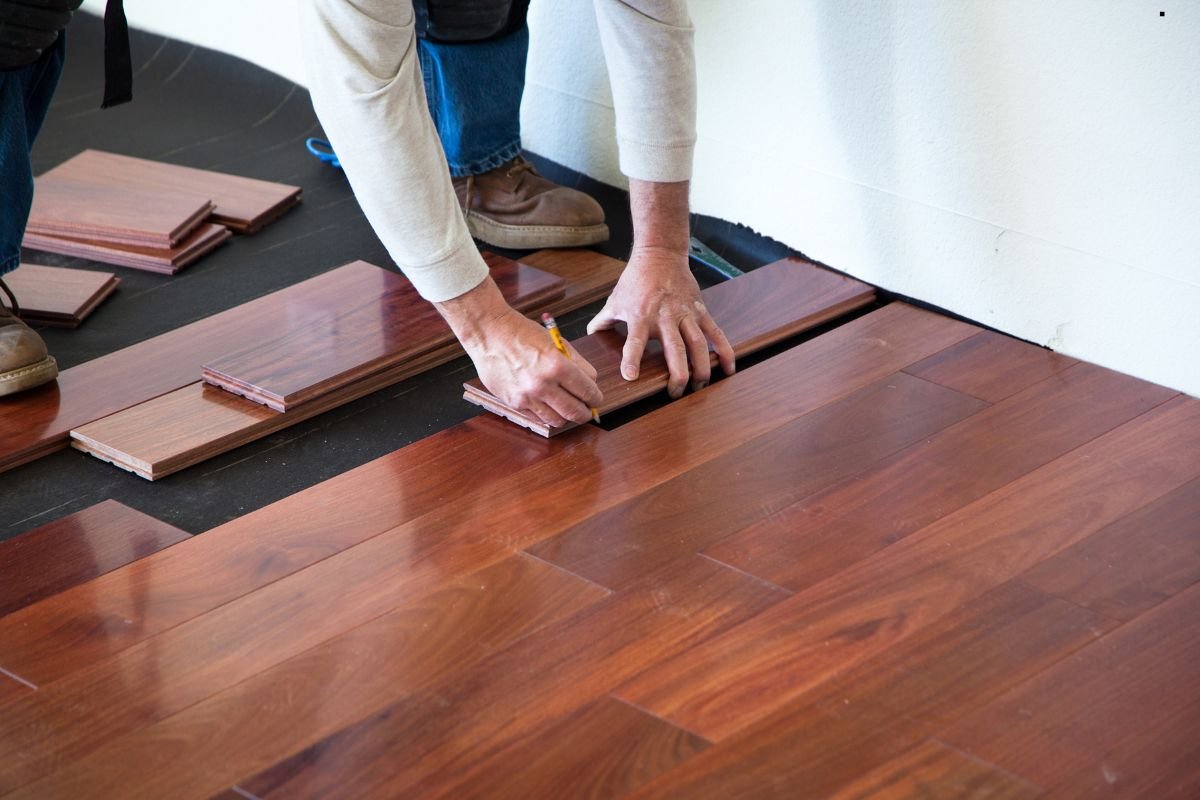 A person marks wooden planks on the floor, preparing for installation, surrounded by laid-out flooring pieces.