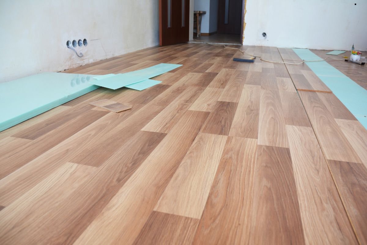 A newly installed wooden floor with light and dark planks, partially covered by green insulation sheets, in a bare room.