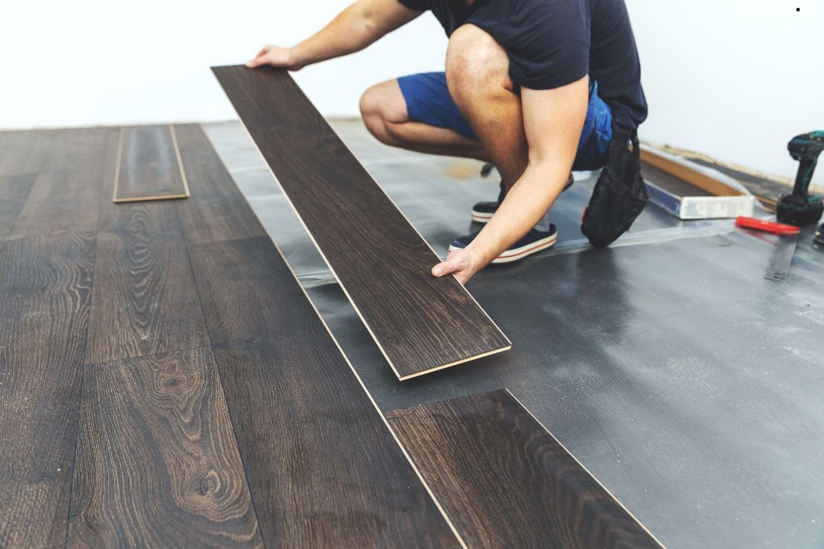 A man in a navy shirt and shorts is installing dark wooden floor planks in a room, carefully placing one on the ground.