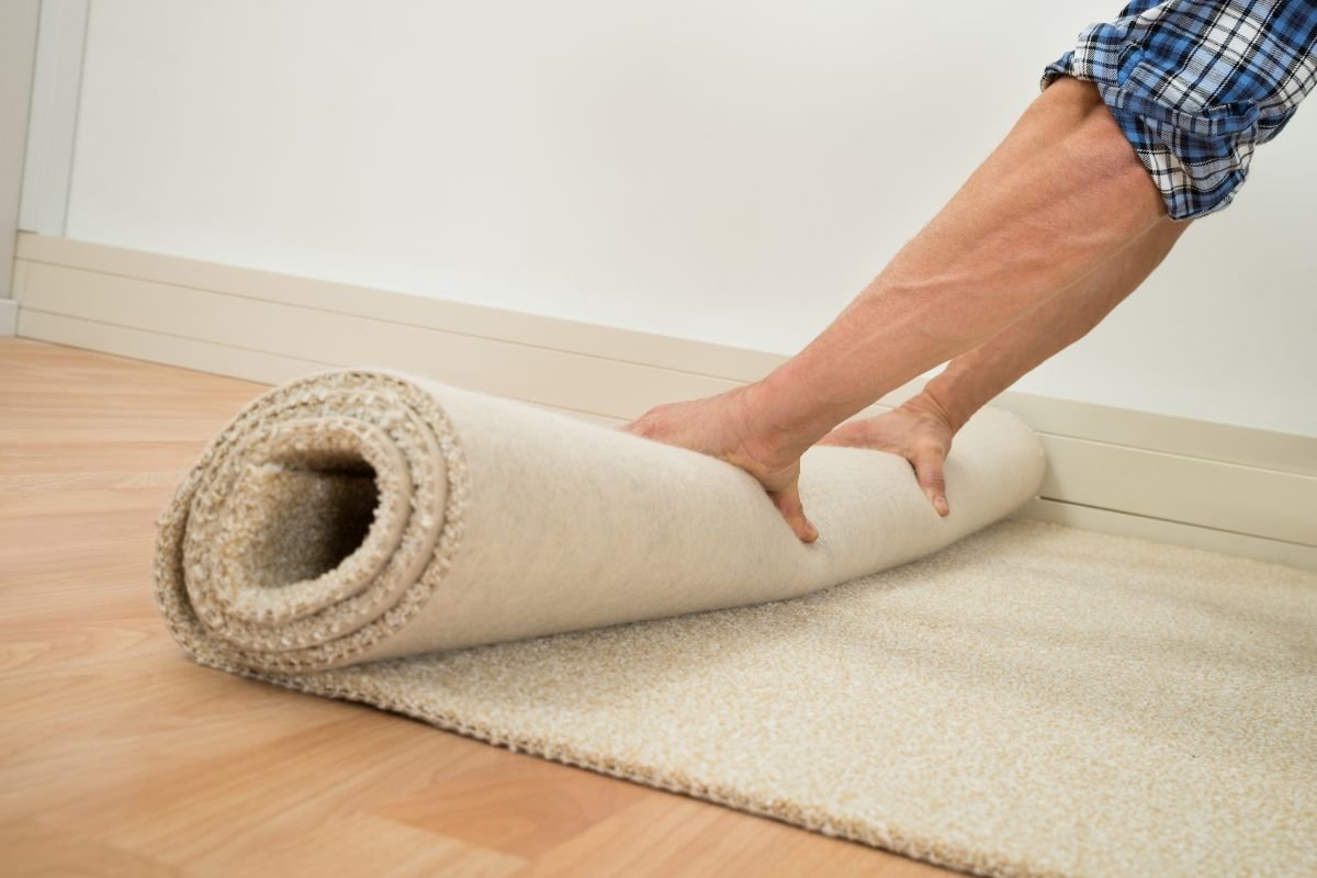 A person rolls out a light beige carpet on a wooden floor, preparing it for placement in a room with white walls.